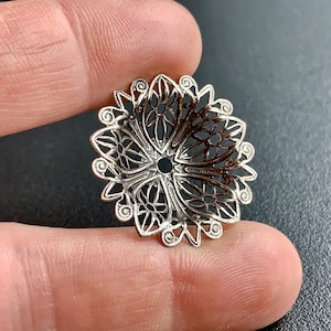 Puede incluir: Adorno de metal plateado filigranado con forma circular y un delicado diseño floral. El adorno tiene un agujero en el centro para sujetarlo a un proyecto.