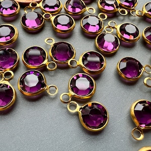 Puede incluir: Un grupo de cuentas de vidrio morado con engastes de metal dorado. Cada cuenta tiene un bucle a cada lado para sujetar a joyas u otras manualidades.