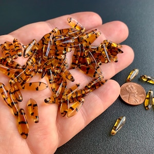 May include: A pile of brown and black glass beads with a tiger stripe pattern. The beads are about 1 inch long and are scattered on a hand. A penny is in the background for size reference.