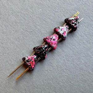 Puede incluir: Cinco cuentas de vidrio triangulares con un patrón moteado negro, rosa y plateado enhebradas en un alambre plateado.