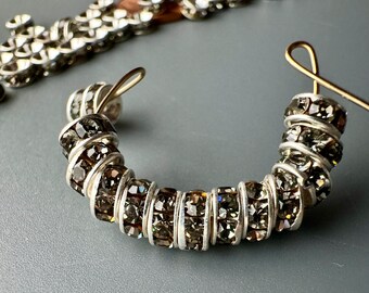 May include: A silver hoop earring with a row of dark gray rhinestones. The earring is open and ready to be worn.