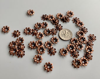 Puede incluir: Una colección de cuentas de metal de color cobre en forma de flor. Las cuentas tienen un diseño en relieve y están esparcidas sobre una superficie blanca.