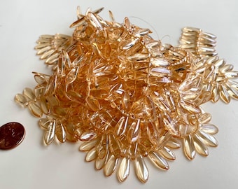 May include: A collection of amber-colored, leaf-shaped beads. The beads are clustered together, with a penny for size comparison. The beads have a translucent quality, and are likely made of glass or acrylic. The beads are arranged in a floral pattern.