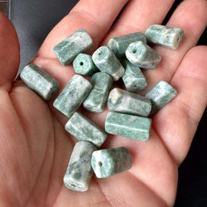 May include: A collection of green and white stone beads. The beads are cylindrical with a hexagonal shape and a hole drilled through the center. The beads are held in a hand.