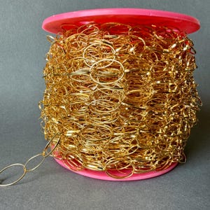 Puede incluir: Un carrete de cadena de color dorado con eslabones ovalados. La cadena está enrollada alrededor de un carrete de plástico rosa.