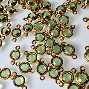 Puede incluir: Una colección de piedras de vidrio facetadas de color verde claro engastadas en biseles de metal con tono de latón antiguo. Cada piedra presenta dos pequeños bucles circulares en los lados opuestos para conectar componentes de joyería.