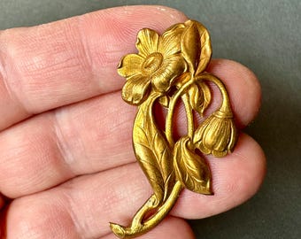 May include: A gold-toned floral brooch featuring a detailed design of a flower with multiple petals, two buds, and leaves. The brooch has a vintage aesthetic and is likely made of metal. The intricate details and warm color suggest a classic jewelry piece.