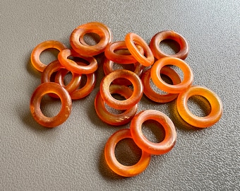 Op de afbeelding: Een groep van 14 amberkleurige ringen, sommige met een lichte oranje tint, liggen op een grijs oppervlak. De ringen zijn allemaal glad en rond en lijken gemaakt te zijn van een natuurlijk materiaal.