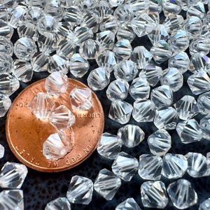 Puede incluir: Primer plano de una gran cantidad de cuentas de vidrio transparentes. Las cuentas son facetadas y tienen un ligero brillo iridiscente. Se incluye una moneda de un céntimo de euro para la escala.
