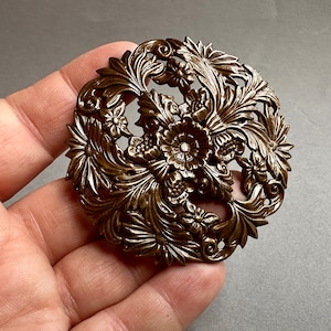 Op de afbeelding: Een grote, sierlijke, goudkleurige metalen broche met een bloemmotief. De broche is ingewikkeld versierd met bladeren en bloemen.