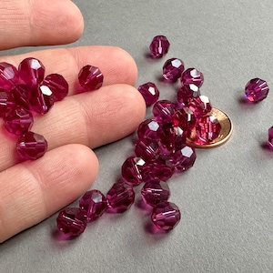 May include: A collection of faceted, deep magenta beads. The beads are scattered on a gray surface, with some held in a hand. A penny is included for size reference. These beads are suitable for jewelry making and crafts.