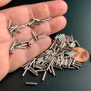 May include: A pile of silver metal tubes, some are arranged in a small pile on a hand, and the rest are scattered on a black surface. A copper penny is in the pile of tubes.