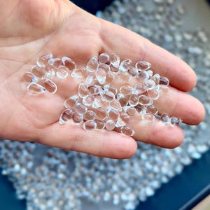 May include: A close-up shot of a hand holding a collection of clear, teardrop-shaped glass beads. The beads are transparent and reflect light, creating a sparkling effect. The background is blurred, with a hint of other similar beads.