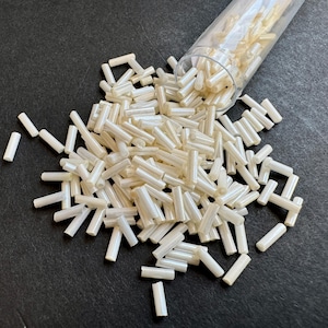 Op de afbeelding: Een close-up van een glazen pot gevuld met witte, iriserende glazen kralen. De kralen zijn cilindrisch van vorm en hebben een gladde, glanzende afwerking.