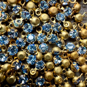 May include: Close-up of a collection of gold-coloured metal components with light blue, round rhinestones. The rhinestones are set in a pronged setting, and the metal components have small loops for attachment. The image is a macro shot.