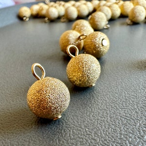 May include: A close-up of two gold-colored, textured, round ornaments with a loop at the top. The ornaments are on a gray, textured surface.