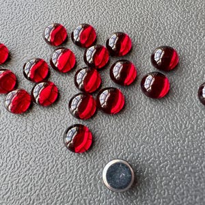May include: A group of 17 red glass cabochons, some with a darker red hue, scattered on a grey textured surface.