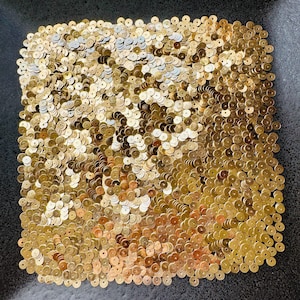 Può includere: Primo piano di un quadrato di paillettes dorate. Le paillettes, piccole e rotonde, sono fittamente impacchettate, creando una superficie strutturata e scintillante. Il colore varia dall'oro brillante a un oro più tenue, con uno sfondo grigio scuro.