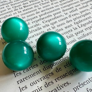 May include: Four round, green glass beads with a glossy finish. The beads are arranged on a page of a book.