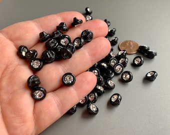 May include: A large pile of black buttons with clear rhinestone centers. The buttons are scattered on a hand and a gray surface.