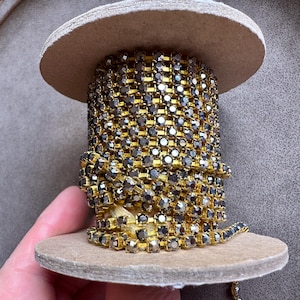 May include: A spool of gold-toned rhinestone chain. The chain is wrapped around a cardboard spool. The rhinestones are a dark, smoky color, and the metal is a bright gold. The spool is sitting on a neutral background.