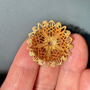 Puede incluir: Pendiente de flor de filigrana dorada con un diseño delicado e intrincado. El pendiente es redondo y tiene un agujero en el centro para sujetar una cadena o cordón.