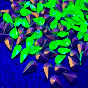 Puede incluir: Una colección de gemas facetadas en forma de lágrima en vibrantes tonos verde neón y púrpura intenso. Las gemas tienen una base de color oro metálico y están dispersas sobre una superficie azul oscuro, creando un contraste llamativo.