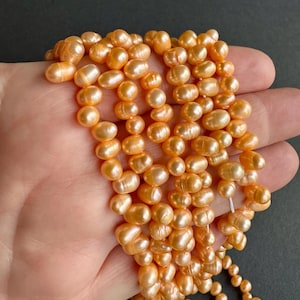 May include: A strand of peach-coloured freshwater pearls. The pearls are irregular in shape and have a slightly iridescent sheen.