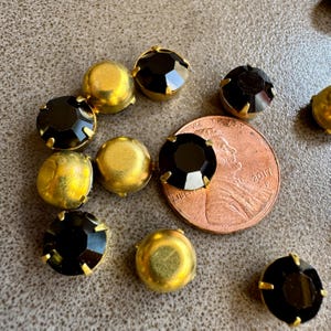 May include: A close-up of black and gold rhinestones. The rhinestones are round and have a faceted design. They are arranged in a random pattern on a brown surface.  The rhinestones are about the size of a US penny.