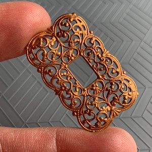 May include: A gold-toned metal filigree embellishment with a rectangular cutout in the center. The intricate design features a repeating pattern of swirls and loops.