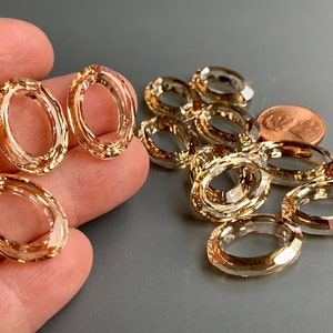 May include: A group of gold-toned oval-shaped crystal beads. The beads are faceted and have a clear, slightly amber hue. The beads are arranged in a loose pile on a gray surface. A penny is included for scale.