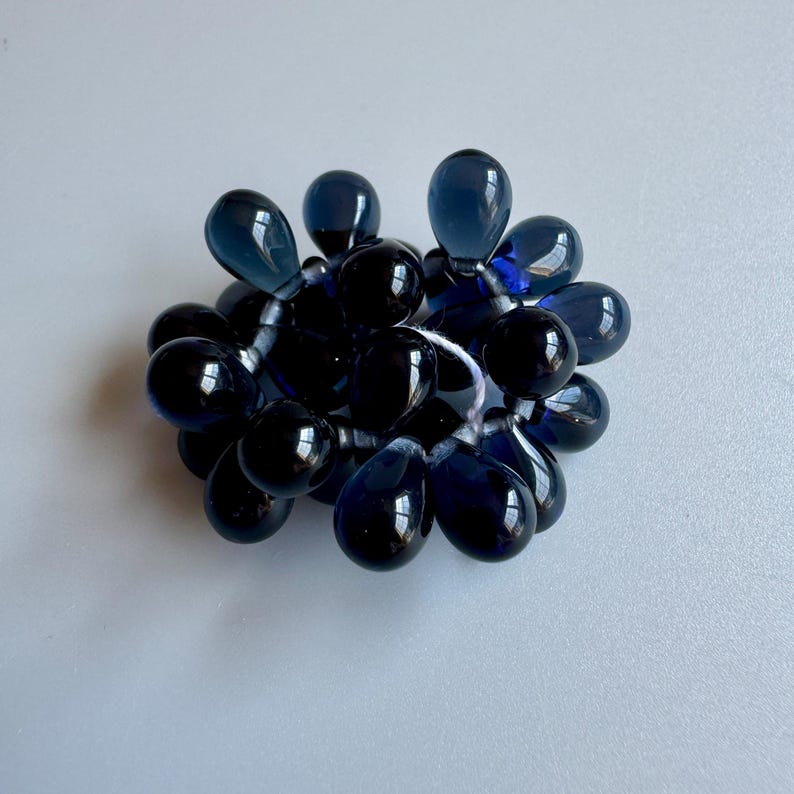 May include: A cluster of dark blue glass beads in the shape of teardrops. The beads are strung together on a white thread.