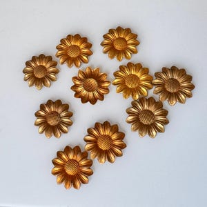 May include: A collection of ten golden sunflower-shaped embellishments. Each flower has detailed petals and a textured center. The metallic finish gives them a vintage or antique appearance, suitable for craft projects or decorative purposes.