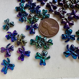 May include: Iridescent shamrock-shaped beads in shades of blue, purple, and green are scattered across a light surface. A US penny is included for size reference. These beads are ideal for jewelry making and craft projects.