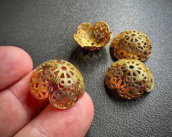 Puede incluir: Cuatro tapas de cuentas de metal de color dorado con intrincados diseños de filigrana. Las tapas son redondas con un fondo plano y una parte superior abovedada. Tienen un delicado patrón calado.