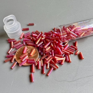 Puede incluir: Pequeñas cuentas de vidrio rojas e iridiscentes, dispersas sobre una superficie gris. Las cuentas cilíndricas se derraman de un tubo de plástico transparente. Una moneda está al lado de las cuentas para la comparación de tamaño.