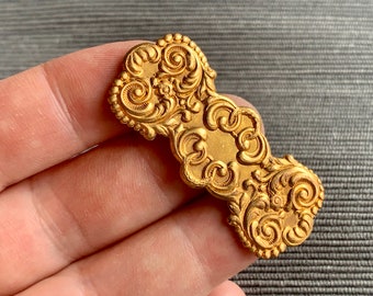 May include: A gold-toned metal decorative embellishment with a detailed floral design. The embellishment has a long, narrow shape with intricate swirls and curves.