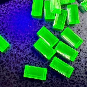 May include: Bright neon green rectangular gemstones scattered on a dark blue surface. The gemstones have a polished, faceted appearance, catching the light. The overall effect is a vibrant display of color and texture, suggesting a collection of precious stones.