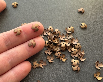 Puede incluir: Un montón de pequeños amuletos de metal de color dorado con forma de estrellas de tres puntas. Los amuletos tienen una superficie texturizada y un agujero en el centro.
