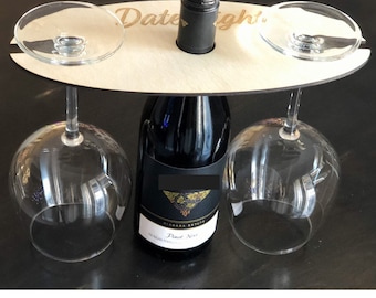 May include: Wooden wine bottle and glass holder with the text "Date Night" engraved on it. The holder has two slots for wine glasses and a center slot for a bottle of wine.
