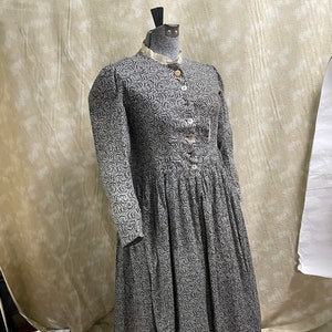 Puede incluir: Un vestido de manga larga con estampado floral en tonos blanco y negro. El vestido tiene un cuello alto y cierre de botones en la parte delantera. Está hecho de una tela ligera y tiene una falda fluida.