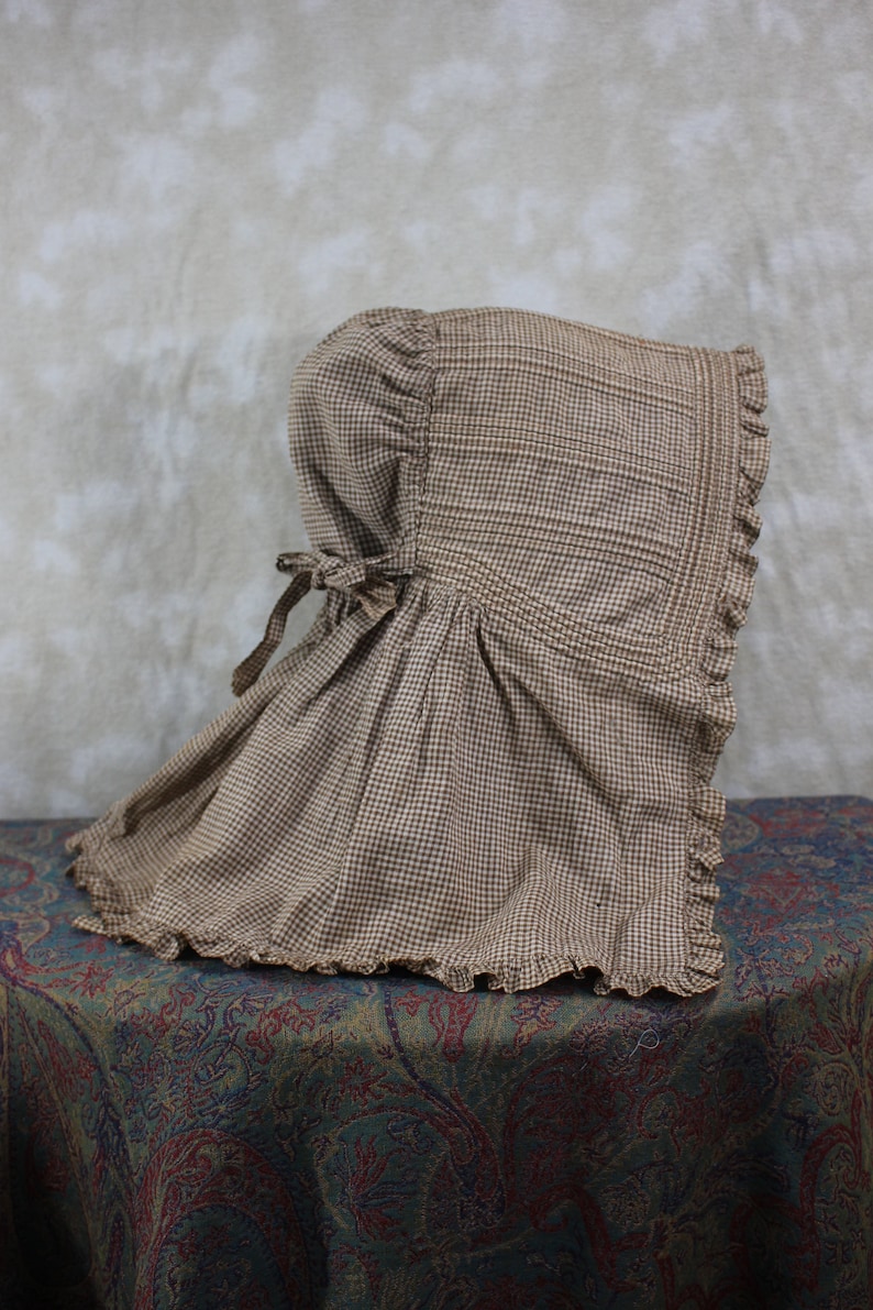 May include: A brown and white gingham bonnet with a ruffled edge. The bonnet is tied at the back with a brown ribbon.