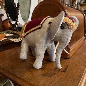 Può includere: Un giocattolo di elefante di peluche grigio con una sella bordeaux e dorata. L'elefante ha zanne bianche e occhi neri. Il giocattolo è seduto su un tavolo di legno.