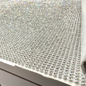 May include: A close-up of a sheet of silver-toned rhinestones, arranged in a grid pattern. The rhinestones are small and have a reflective surface.