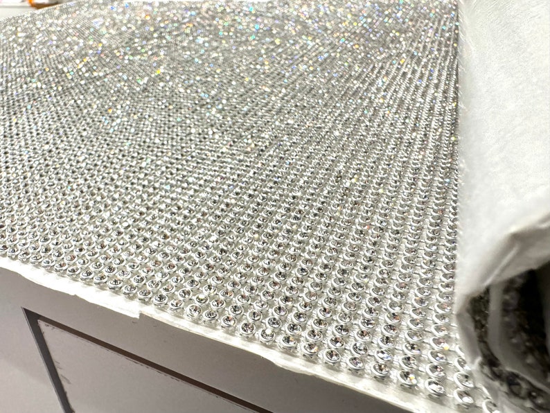 May include: A close-up of a sheet of silver-toned rhinestones, arranged in a grid pattern. The rhinestones are small and have a reflective surface.