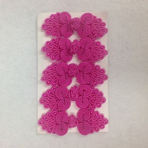 Könnte beinhalten: Sechs Paare rosa Stoffknöpfe mit chinesischem Knotendesign und Perlenverzierung. Die Knöpfe sind in zwei Reihen zu je drei auf einer weißen Karte angeordnet.