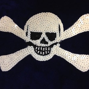 Puede incluir: Un diseño de calavera y huesos cruzados, adornado con lentejuelas blancas, sobre un fondo azul oscuro. La calavera presenta cuencas de ojos y boca negras, también con detalles negros. Los huesos cruzados están colocados detrás de la calavera.