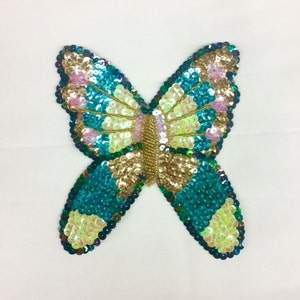May include: A sequined butterfly applique in shades of teal, gold, pink, and light green. The butterfly has a beaded body and is set against a white background. The sequins are arranged in a pattern, creating a shimmering effect.