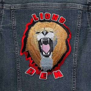 May include: A denim jacket with an embroidered patch featuring a roaring lion with the words "Lions Den" in red.