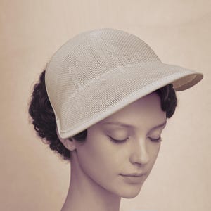 May include: A cream-colored, mesh-knit hat with a small brim. The hat has a rounded crown and a structured design. The hat is worn by a person with dark hair and closed eyes. The background is a soft, neutral tone.